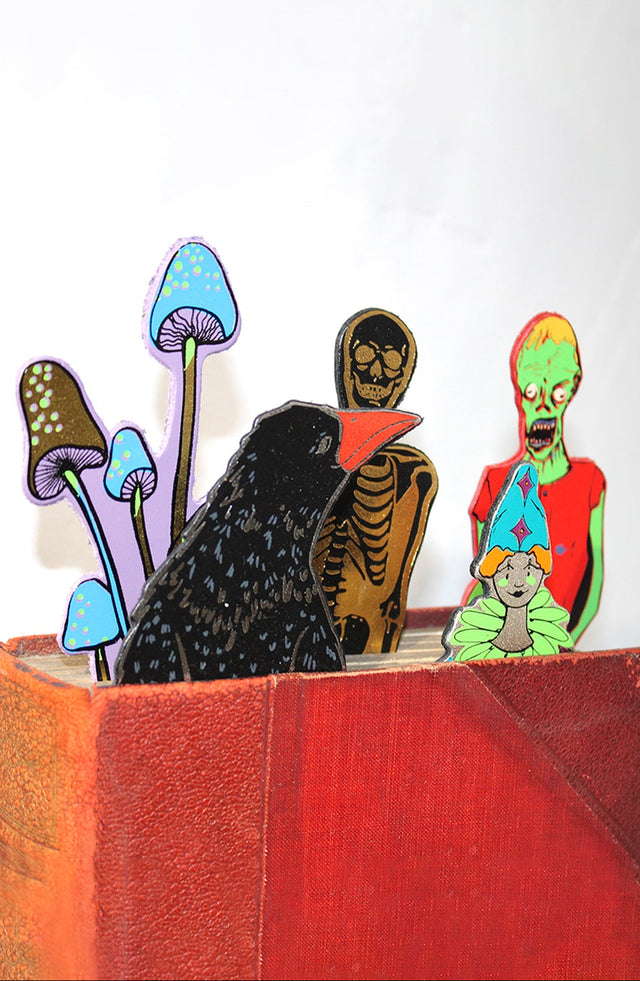 A set of coluorful Halloween-themed leather cutout bookmarks, including a black raven, a skeleton, a green zombie, and blue mushrooms, arranged inside a red-orange book against a plain white background.